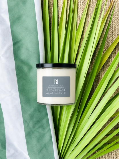Candle labeled 'Beach Day' by Riverhomeco on a leafy background